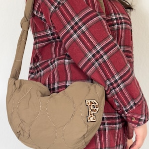 May include: A brown quilted heart-shaped crossbody bag with a leopard print "P" patch. The bag has a long adjustable strap and is worn over a red and black checked shirt.