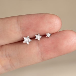 Pode incluir: Três brincos prateados em forma de estrela de tamanhos variados. Os brincos são cravejados com pedras preciosas claras e brilhantes. O brinco menor está à direita, o maior à esquerda. Os brincos são exibidos em um fundo claro.