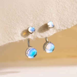 Könnte beinhalten: Silberne Ohrringe mit schillernden Mondstein-Tropfen. Die Ohrringe haben einen Stecker und einen größeren, baumelnden Mondstein. Die Mondsteine zeigen Blau- und Weißtöne mit einem subtilen Glanz. Präsentiert auf cremefarbenem Hintergrund.
