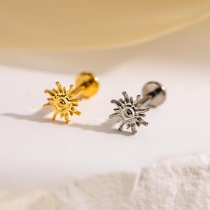 May include: Two sun-shaped ear studs. One is gold-colored, the other is silver-colored. Each stud has a small, round, flat back and a decorative sunburst design on the front. The studs are displayed on a white surface.