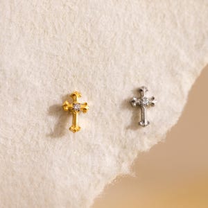 May include: Two small cross-shaped earrings. One is gold-colored with a central clear stone, the other is silver-colored with a clear stone. The earrings are displayed on a white, textured surface.