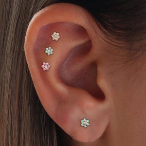 Pode incluir: Close-up de uma orelha adornada com quatro brincos pequenos em forma de flor. Os brincos apresentam uma gema central rodeada por pétalas em tons de rosa, azul e verde. Os brincos são em metal dourado.