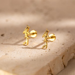 May include: A pair of gold-coloured stud earrings. Each earring features a detailed koi fish design. The earrings are set against a neutral, textured background, highlighting their metallic sheen and intricate form.