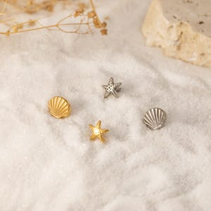 Puede incluir: Cuatro pequeños abalorios con temática de playa están dispersos sobre una superficie blanca. Los abalorios incluyen dos estrellas de mar, una dorada y otra plateada, y dos conchas, una dorada y otra plateada. Los abalorios son detallados y parecen ser de metal.