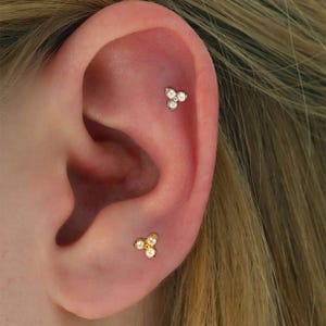 Puede incluir: Primer plano de una oreja con dos joyas. La joya superior es un pequeño pendiente de plata con tres piedras redondas y transparentes. La joya inferior es un pendiente dorado con tres pequeñas perlas blancas.