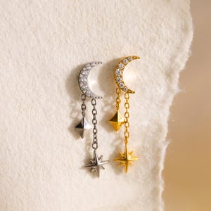 Puede incluir: Un par de pendientes de temática celestial. Uno es plateado con una luna creciente incrustada con piedras transparentes, un dije en forma de diamante y una estrella. El otro es dorado con dijes similares.