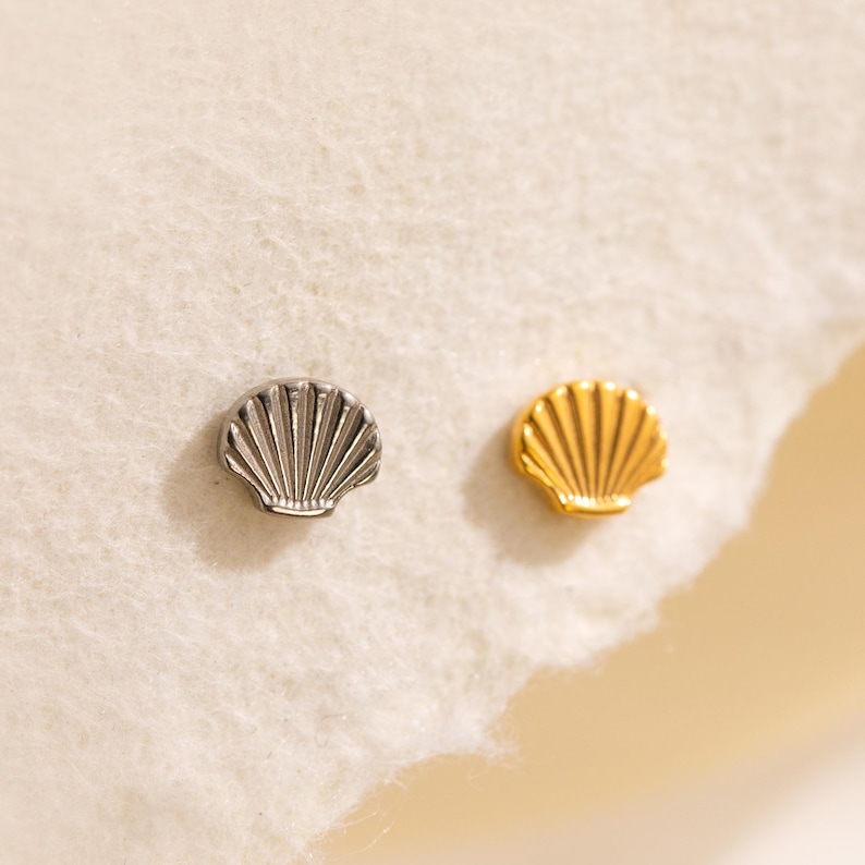 May include: Two small seashell-shaped stud earrings. One is silver-colored, and the other is gold-colored. The earrings are placed on a soft, white surface with a light yellow background.