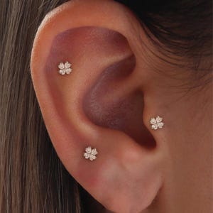 Könnte beinhalten: Nahaufnahme eines Ohrs mit drei kleinen Ohrsteckern in Kleeblattform. Jeder Ohrring hat eine Ansammlung kleiner, funkelnder Steine, wahrscheinlich Zirkonia oder Diamanten, in einem hellen Metall.