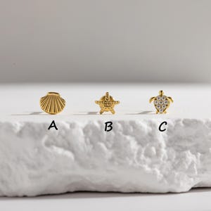May include: Three gold-colored stud earrings in a row. The earrings are shaped like a seashell, a starfish, and a turtle. The turtle earring has a clear stone design. The letters A, B, and C are below each earring.