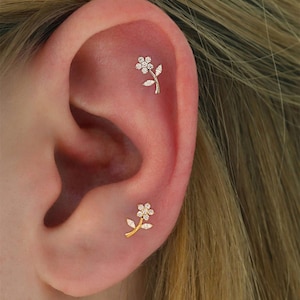 May include: Close-up of an ear with two small flower-shaped earrings. The earrings have a gold stem and leaves, with a cluster of small, clear stones forming the flower. The ear is against a blurred background.