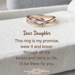 May include: A rose gold ring with a knot design and small diamonds. The ring is in a white box with the text "Dear Daughter This ring is my promise, wear it and know: through all the twists and turns in life, I'll be there for you."