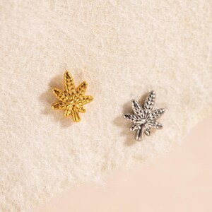 May include: Two small, leaf-shaped earrings. One is gold-colored, the other is silver-colored. The earrings are placed on a light-colored, textured surface.