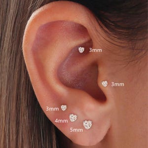 May include: Close-up of an ear with multiple heart-shaped stud earrings. The earrings are 3mm, 4mm, and 5mm in size. The earrings are small, sparkly, and appear to be made of a clear material.