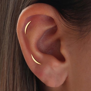 Puede incluir: Primer plano de una oreja con dos pendientes pequeños, curvos y dorados. Los pendientes tienen forma de media luna y están colocados en la parte superior de la oreja. El tono de piel es claro y el cabello es oscuro.