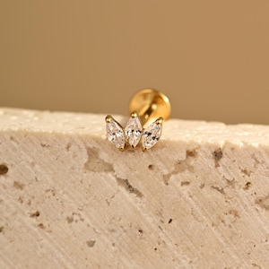 May include: A gold-colored stud earring with three clear, marquise-cut gemstones arranged in a cluster. The earring is set against a textured, light-colored surface, with a soft, neutral background.