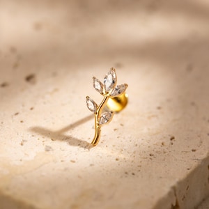 May include: A gold-coloured stud earring shaped like a delicate leaf, featuring five clear, marquise-cut gemstones. The earring is set against a neutral, textured background, with a shadow adding depth.