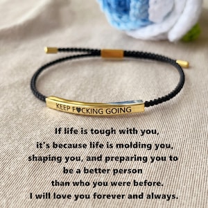 Könnte beinhalten: Ein schwarzes Kordelarmband mit einem goldfarbenen Balken, auf dem die Worte "KEEP FUCKING GOING" eingraviert sind. Das Armband hat goldfarbene Endkappen. Der Hintergrund enthält Text.