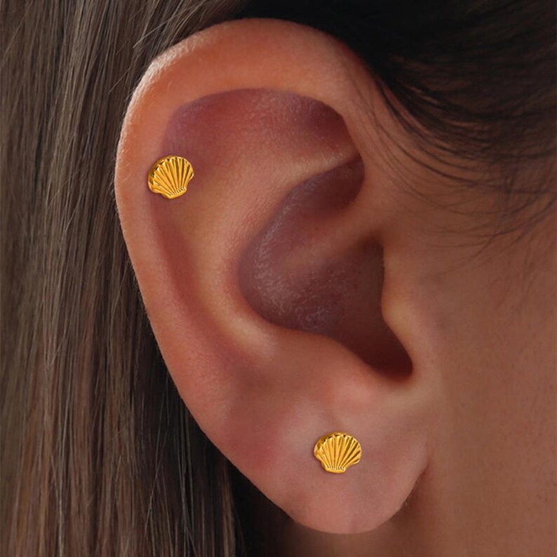 May include: Close-up of a person's ear adorned with two small, gold seashell stud earrings. The earrings are detailed with radiating lines, catching the light. The skin tone is fair, and the hair is a light brown.