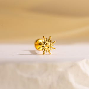 Pode incluir: Um brinco dourado com um design de sol. O brinco apresenta um pequeno pino redondo e uma forma de sol detalhada com raios radiantes. O design do sol é centrado no brinco, com um acabamento liso e polido.