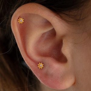 May include: Close-up of an ear with two small, gold-colored stud earrings. Each earring has a sunburst design with a small, clear stone in the center. The earrings are set against the skin of the ear.