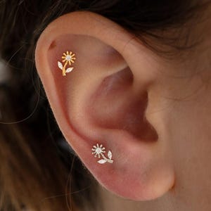Può includere: Due orecchini a forma di fiore color oro, ciascuno con una pietra centrale trasparente e delicati accenti di foglie, sono esposti su un orecchio. Gli orecchini sono piccoli e dettagliati, mostrando un design floreale. L'orecchio è in soft focus, evidenziando i gioielli.