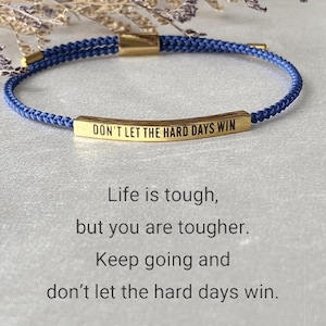 Könnte beinhalten: Ein blaues Kordelarmband mit einem goldenen Barren-Anhänger, auf dem "DON'T LET THE HARD DAYS WIN" steht. Das Armband ist eine Erinnerung, positiv zu bleiben und weiterzumachen.