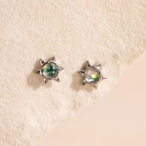 May include: Two star-shaped stud earrings. One earring features a green and white stone, while the other has an iridescent, multi-colored stone. The earrings have a silver-colored metal setting and are displayed on a light-colored, textured surface.