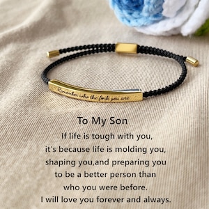 Op de afbeelding: Een zwarte gevlochten armband met een gouden staafbedel gegraveerd met de tekst "Remember who the fuck you are". De armband wordt vergezeld van een boodschap die luidt: "To My Son If life is tough with you, it's because life is molding you, shaping you, and preparing you to be a better person than who you were before. I will love you forever and always."