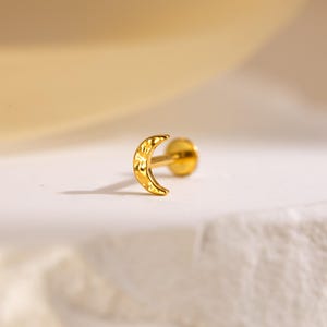 以下が含まれることがあります： 金色の三日月型のイヤリングで、表面はテクスチャ加工されています。イヤリングにはポストと平らな背面があります。イヤリングは軟骨ピアス用に設計されています。イヤリングは白い表面にあります。