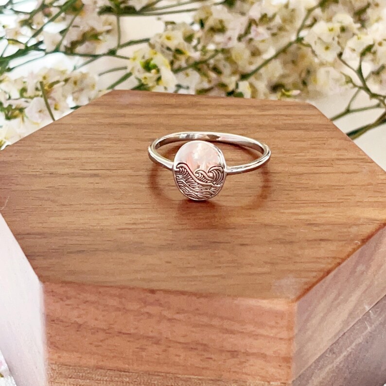 May include: A silver ring with a pink oval-shaped stone featuring an engraved wave design.