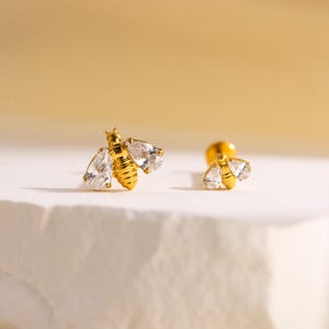 Puede incluir: Dos pendientes dorados con forma de abeja. Cada abeja tiene un cuerpo con detalles acanalados y alas de cristal transparente en forma de pera. Los pendientes están sobre una superficie blanca con un fondo amarillo suave.