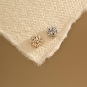 May include: Two snowflake stud earrings. One is gold-toned with a central stone, the other is silver-toned with a central stone. Both earrings are set on a cream-coloured, textured surface.