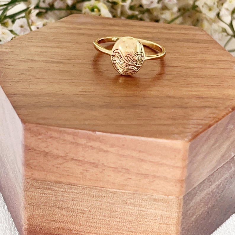 May include: A gold ring featuring a wave design on the top. The ring is displayed on a wooden surface.