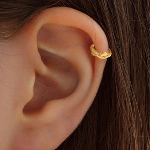 Könnte beinhalten: Ein goldfarbener Ohrklemme mit strukturierter Oberfläche wird am Ohr getragen. Der Ohrklemme ist so konzipiert, dass er den Ohrknorpel umschließt. Der Schmuck hat eine leuchtend goldene Farbe.