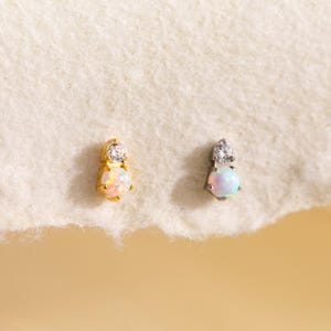 May include: Two stud earrings. One is gold-toned with a round opal and a small clear stone. The other is silver-toned with a similar opal and clear stone. Both earrings are on a white, textured surface.