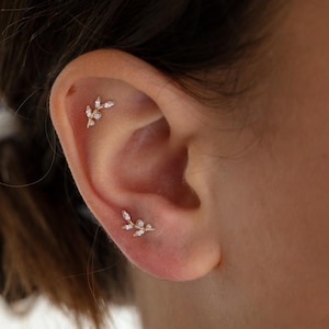 Puede incluir: Primer plano de una oreja adornada con dos pendientes dorados en forma de hoja. Cada pendiente presenta pequeñas gemas claras y brillantes. Los pendientes están colocados en el cartílago y el lóbulo de la oreja.