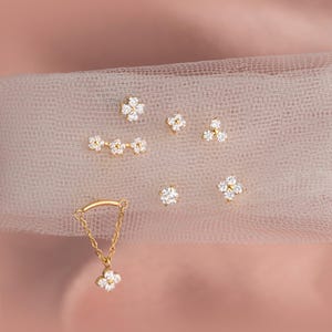 May include: A selection of gold-coloured jewellery pieces with floral designs. The set includes stud earrings, a necklace with a chain, and other small decorative elements. The jewellery is adorned with clear, sparkling gemstones, set against a soft, pink background.