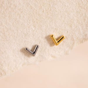 Peut inclure: Deux petites boucles d'oreilles à clous en forme de V. L'une est de couleur argentée, l'autre de couleur dorée. Les boucles d'oreilles sont présentées sur une surface beige clair et texturée.
