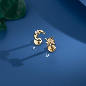 Può includere: Due orecchini a lobo color oro, uno a forma di mezzaluna e l'altro a stella. Entrambi gli orecchini hanno una piccola gemma trasparente. Gli orecchini sono su sfondo blu.