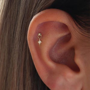 Può includere: Un orecchino color oro con un ciondolo a forma di stella e una piccola gemma trasparente. L'orecchino è attaccato all'orecchio. È un gioiello delicato.