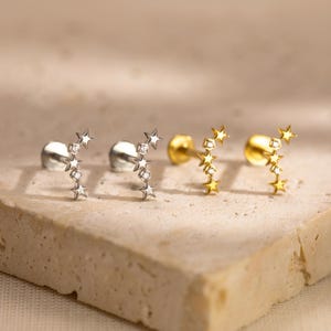 May include: Two pairs of star-shaped earrings. One pair is silver with small clear stones, and the other is gold with small clear stones. The earrings are displayed on a light-colored, textured surface.