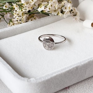May include: A silver ring featuring a round, engraved design of ocean waves. The ring is displayed on a white surface.