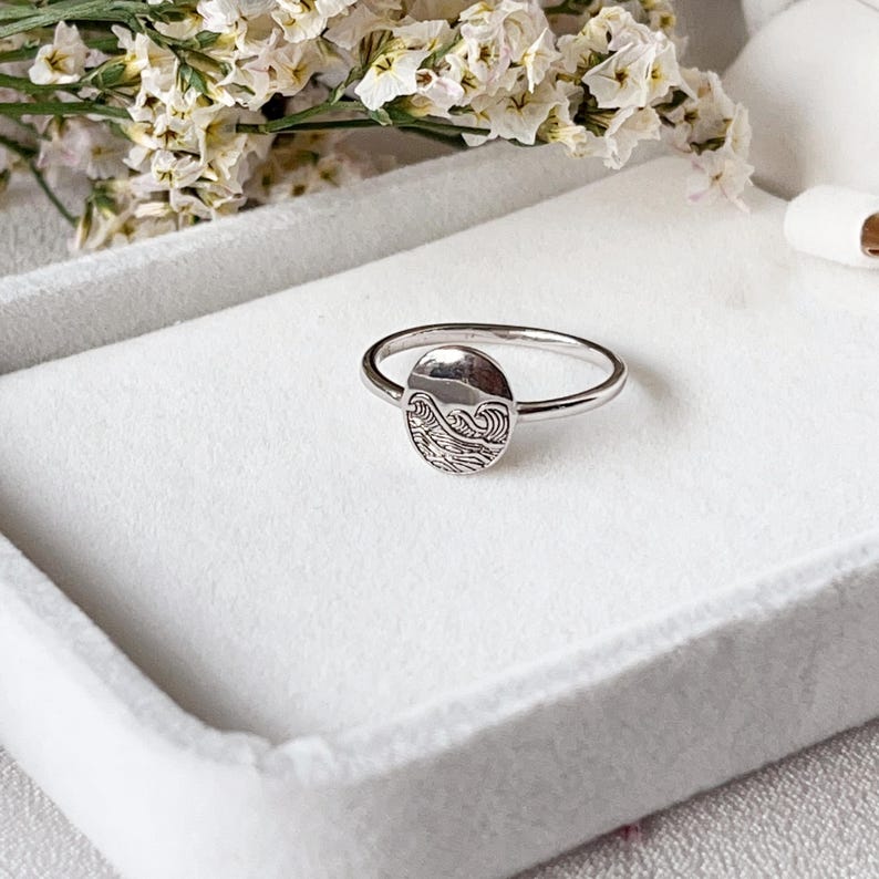 May include: A silver ring featuring a round, engraved design of ocean waves. The ring is displayed on a white surface.