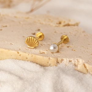 May include: Two gold-colored stud earrings. One earring features a seashell design, and the other has a white pearl. The earrings are displayed on a light-colored, textured surface, with a white, sandy background.