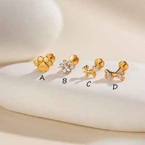 May include: Four gold-colored stud earrings. Designs include a paw print, a flower with clear stones, a dog, and a bone with clear stones. Each earring has a post and backing for secure wear.