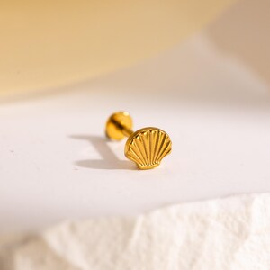 May include: A gold-colored seashell stud earring. The earring features a detailed seashell design on the front and a post on the back. The earring is displayed on a white surface with a soft, yellow background.