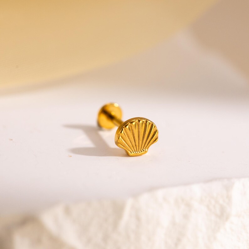 May include: A gold-colored seashell stud earring. The earring features a detailed seashell design on the front and a post on the back. The earring is displayed on a white surface with a soft, yellow background.