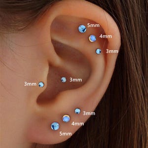 May include: Close-up of an ear with multiple piercings. The ear has several small, light blue stud earrings of varying sizes, ranging from 3mm to 5mm. The earrings are placed in different locations on the ear, showcasing a variety of piercing styles.