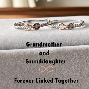 May include: Two infinity rings with small clear stones set in silver bands. The rings are linked together, with one ring featuring a rose gold infinity symbol. The text "Grandmother and Granddaughter Forever Linked Together" is below the rings.