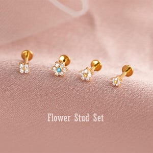May include: A set of four gold-coloured flower stud earrings. Each earring features a flower design with small, clear gemstones. One earring has a blue gemstone in the centre. The text "Flower Stud Set" is below the earrings.